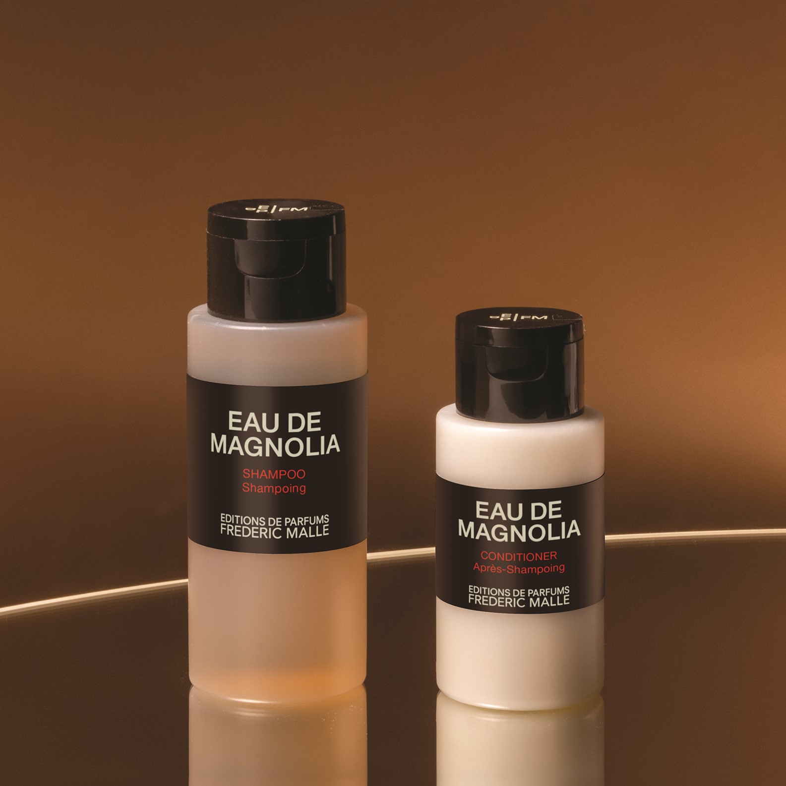 Close-up of Eau de Magnolia shampoo and conditioner bottles by Editions de Parfums Frédéric Malle, displayed against a warm brown background with soft reflections highlighting the frosted bottles and black caps.