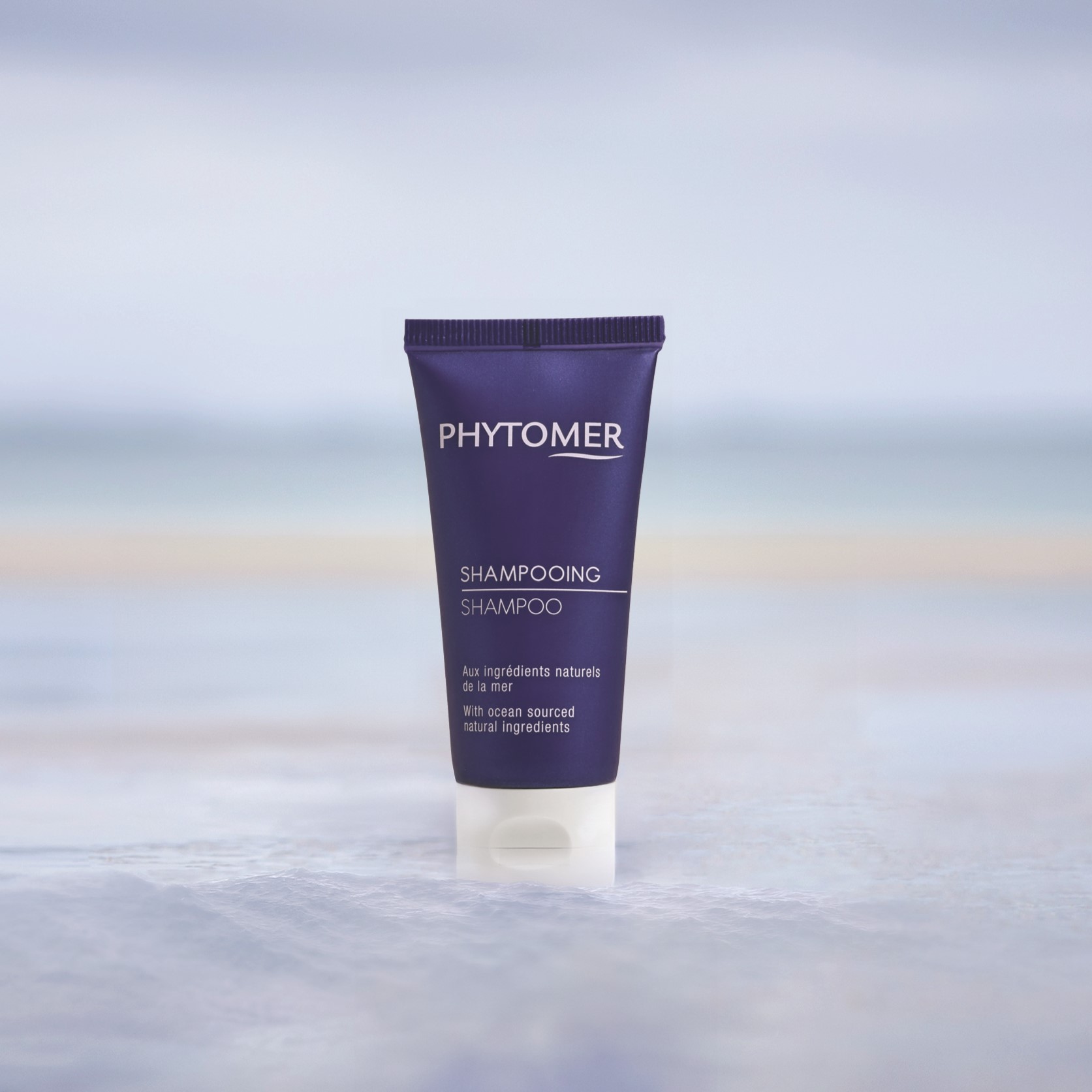 Phytomer tube de shampoing format voyage posé dans de l'eau peu profonde, avec un fond représentant un paysage de bord de mer.