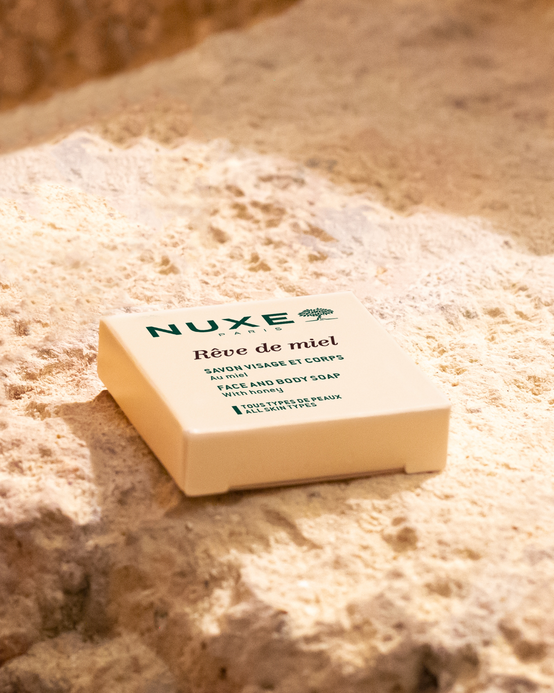 jabón para cara y cuerpo «Rêve de miel» de NUXE, jabón sobre una superficie arenosa con textura, resaltado por una cálida luz natural que realza el envase de color beige y el texto verde.