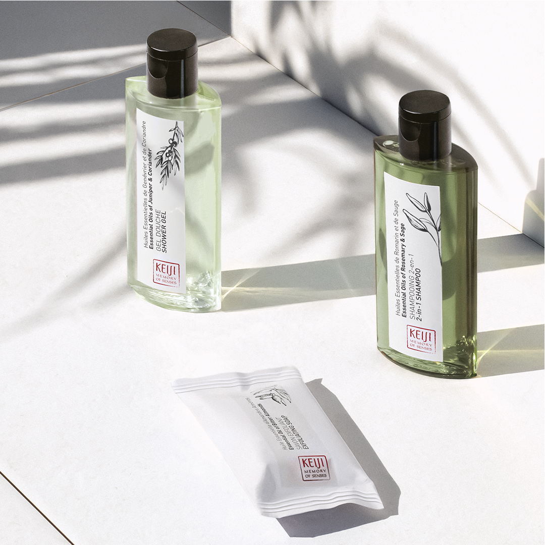 Keiji Memory of Senses bathroom amenities arranged on a sunlit white surface, including two green-toned bottles with black caps and a sealed white sachet in the foreground, with soft plant shadows creating a natural, minimalist atmosphere.