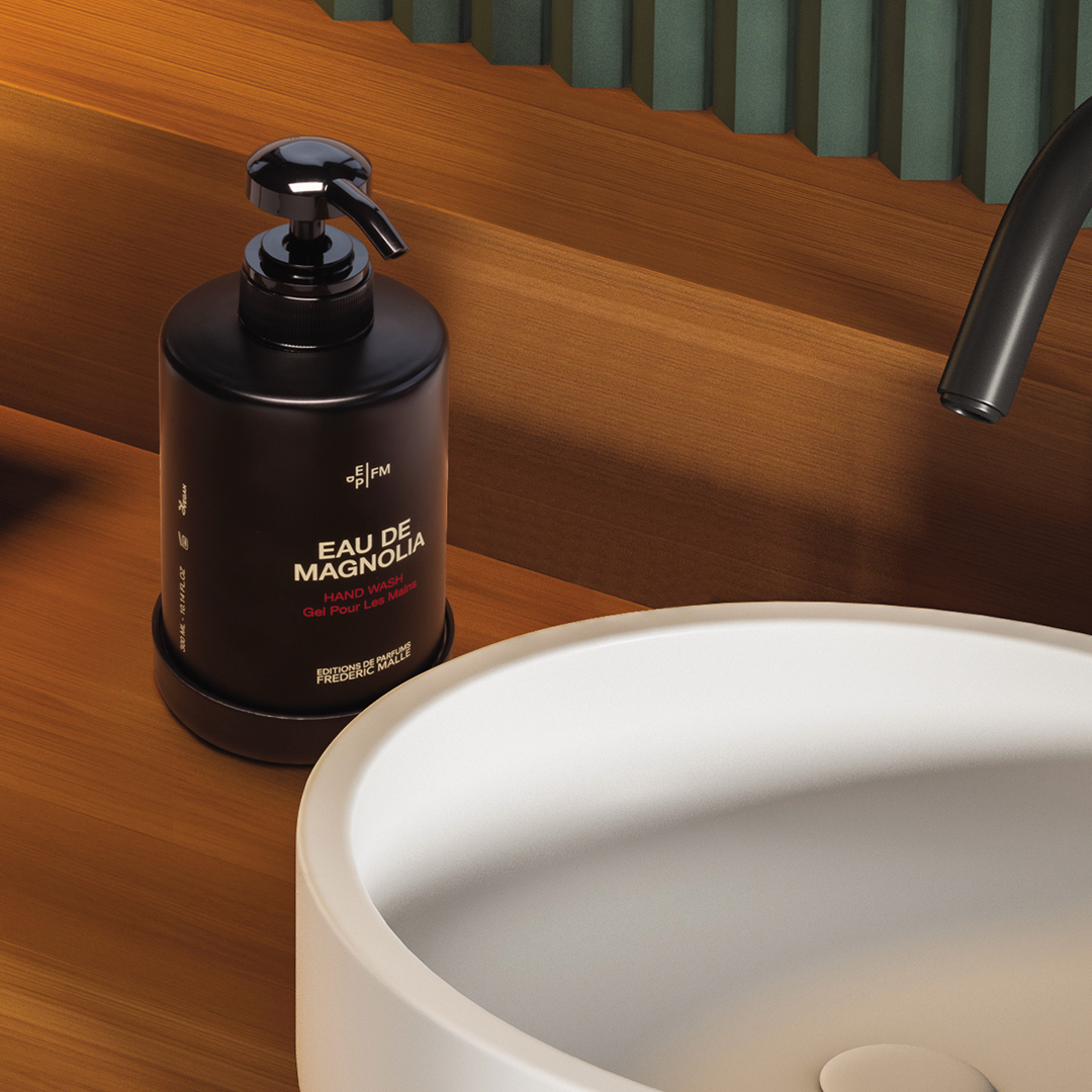 Eau de Magnolia hand wash bottle with a black pump placed next to a modern white sink on a warm wooden countertop, with a curved black faucet and textured green wall detail in the background.