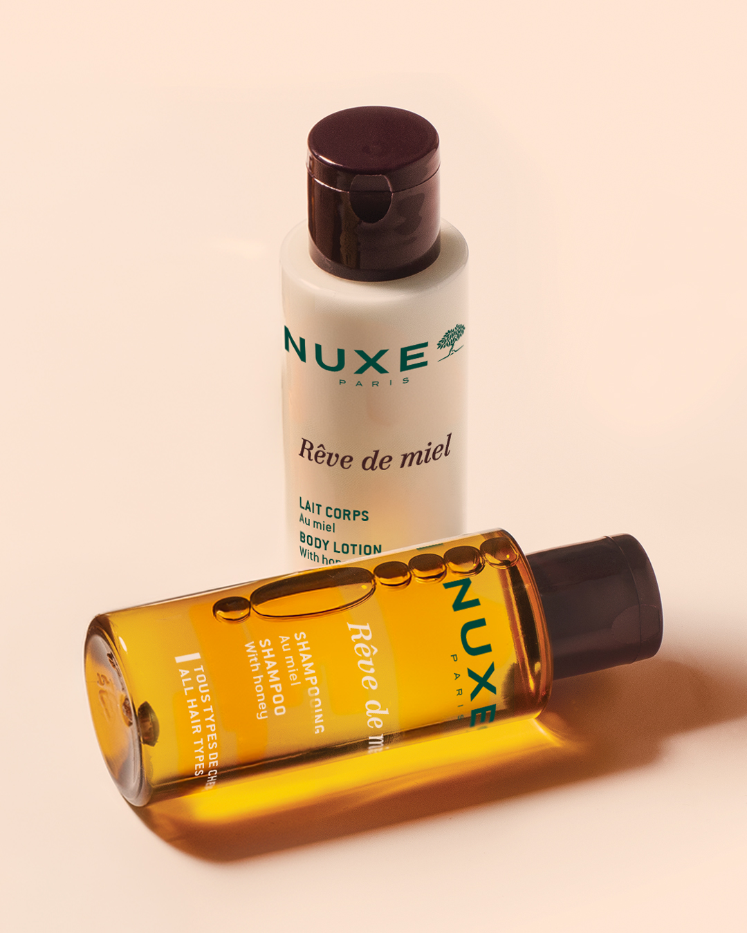 Productos de la gama «Rêve de miel» de NUXE Paris sobre un fondo beige: una loción corporal de miel en posición vertical y una botellas de champú de color ámbar botellas horizontal, con cálidos reflejos de luz.