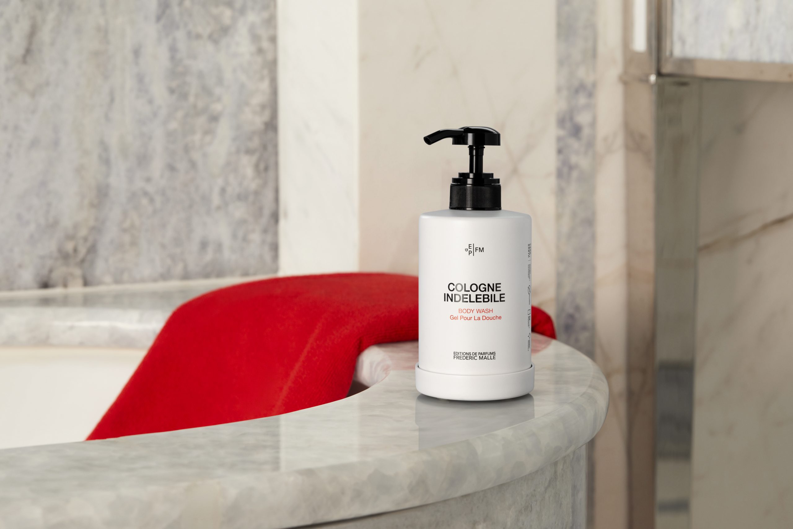 Cologne Indélébile body wash bottle placed on a marble countertop beside a bathtub, with a red towel draped in the background, creating a warm and elegant bathroom setting.