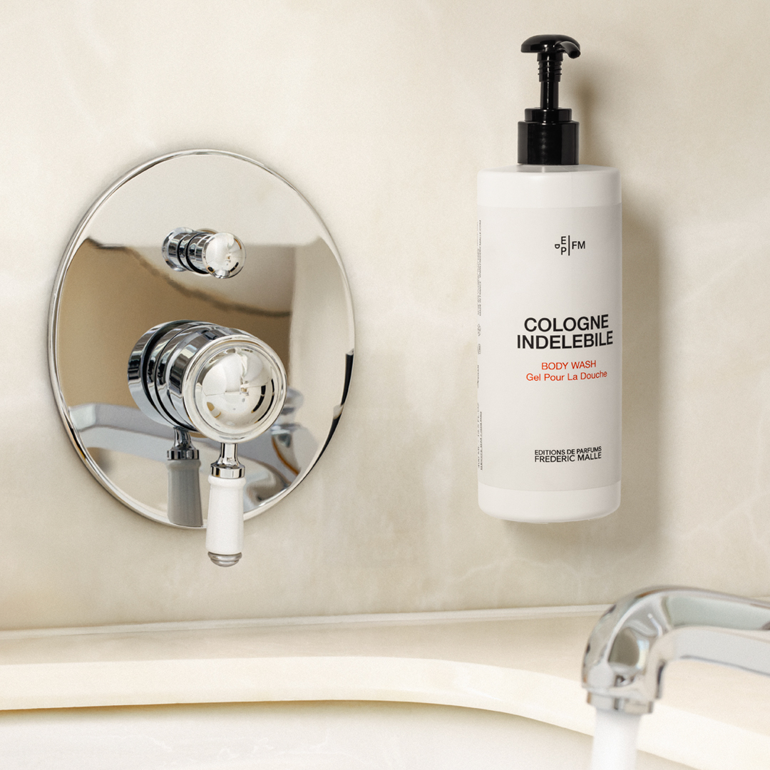 Wall-mounted Cologne Indélébile body wash bottle by Editions de Parfums Frédéric Malle next to a round chrome shower control, set in a clean beige-toned bathroom with a minimalist aesthetic.