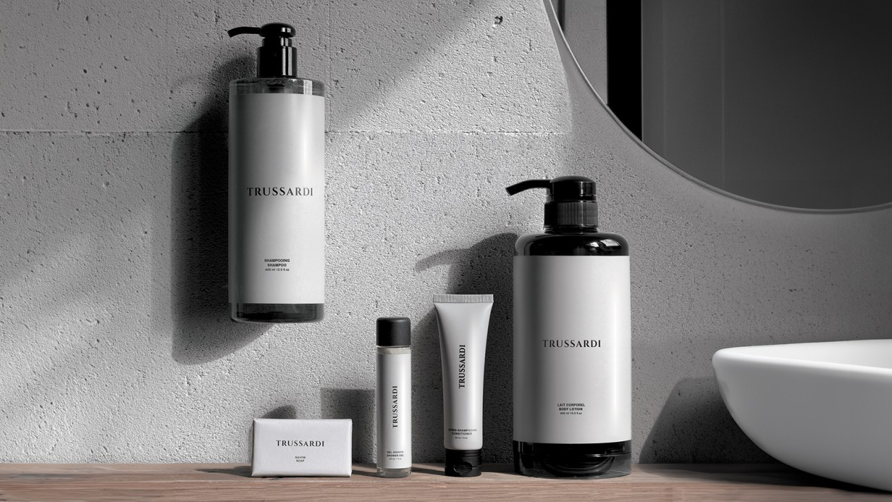 Trussardi bath and body product lineup displayed on a wooden countertop in a minimalist bathroom setting, including a wall-mounted shampoo bottle, body lotion pump, small tubes, and a boxed soap next to a white sink and round mirror.