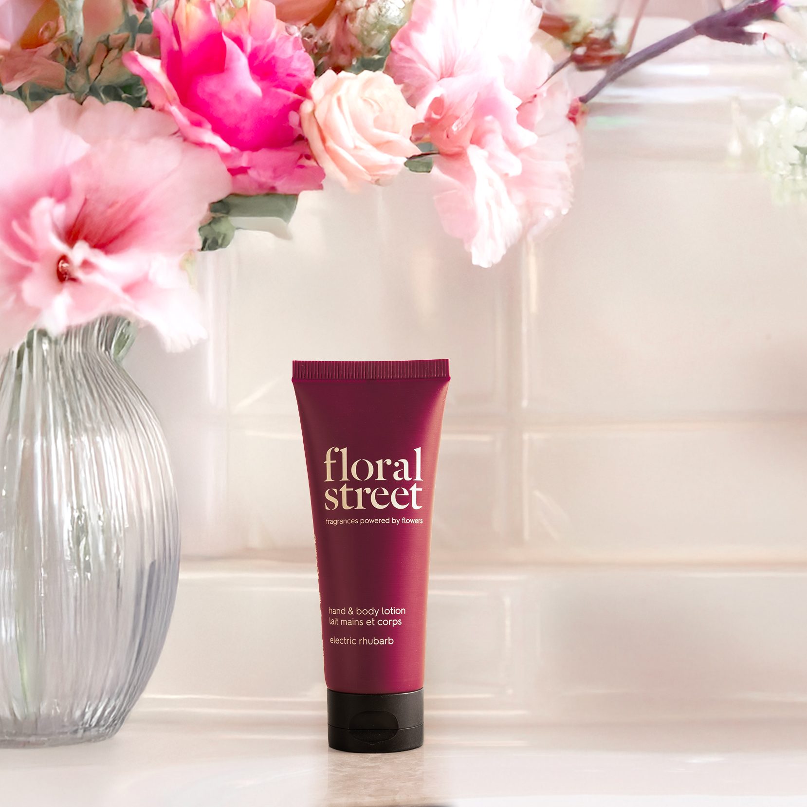 Floral Street Electric Rhubarb hand and body lotion tube placed beside a glass vase of pink and white flowers in a soft, bright interior setting.