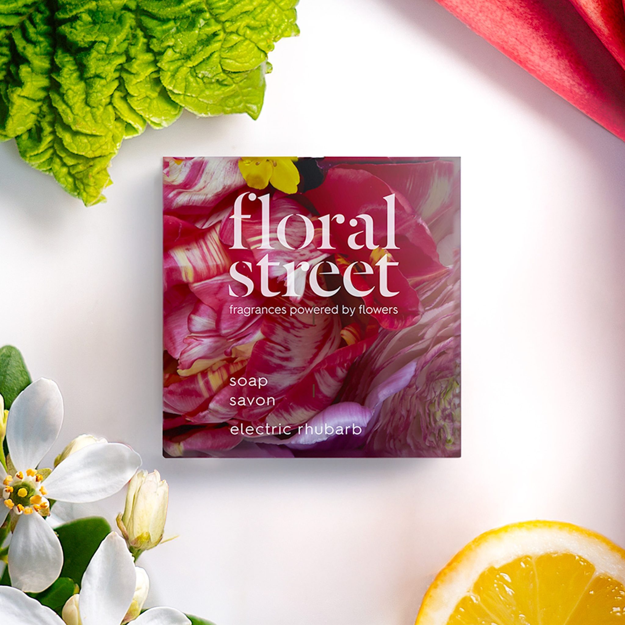 Floral Street Electric Rhubarb soap bar packaging surrounded by fresh rhubarb stalks, white flowers, green leaves, and lemon slice on a clean white background.