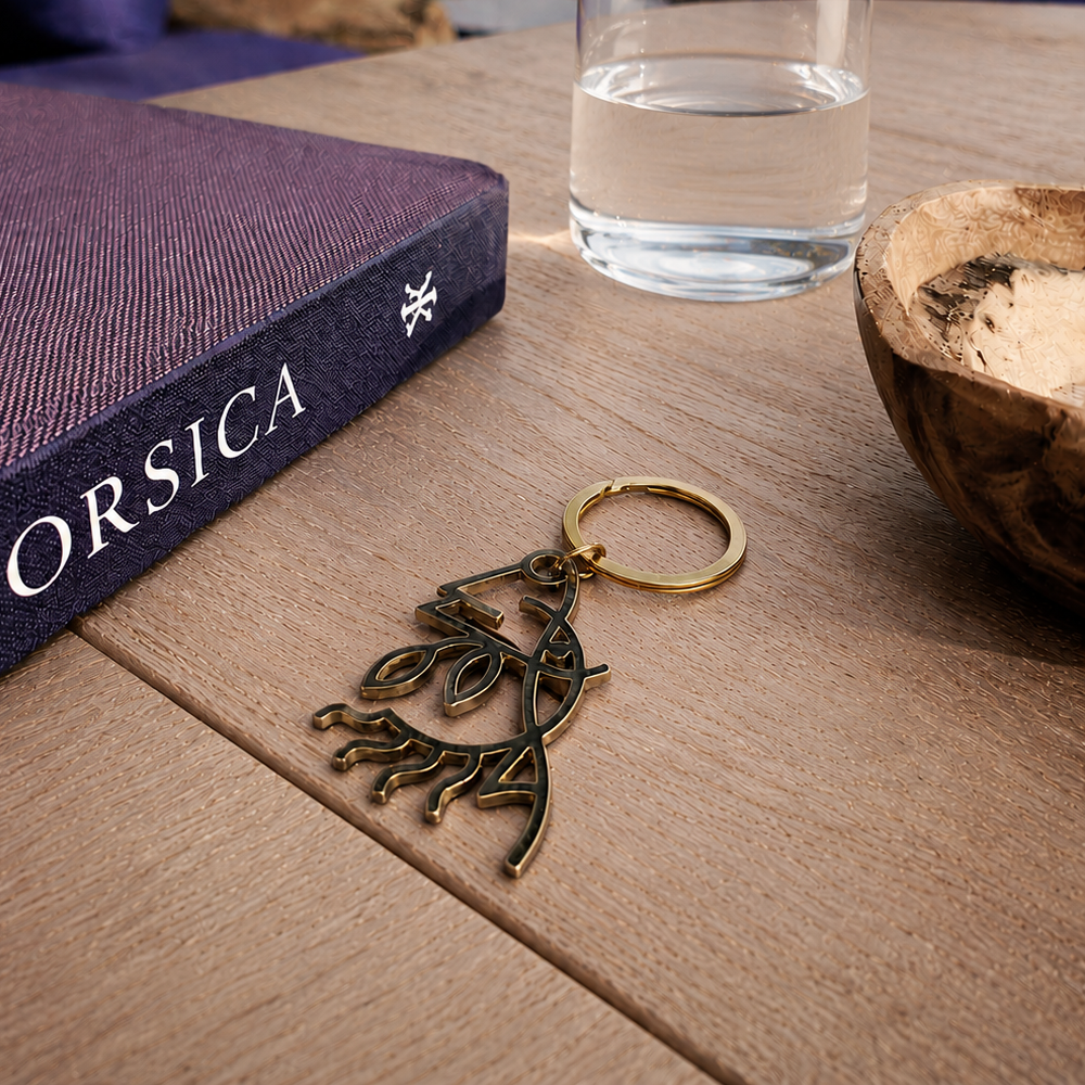 Branded Domaine de Murtoli metal keychain displayed on a wooden table beside a glass of water and a presentation box, with a blurred seaside terrace in the background