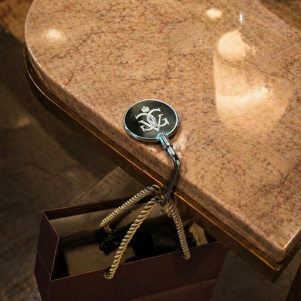 A polished granite tabletop with rounded edges is shown in a warm indoor setting. A metal handbag hook engraved with an elegant monogram is attached to the table edge, suspending a dark gift bag with braided rope handles. Reflections from overhead lighting are visible on the glossy stone surface, highlighting the practical use of the table-mounted bag holder.