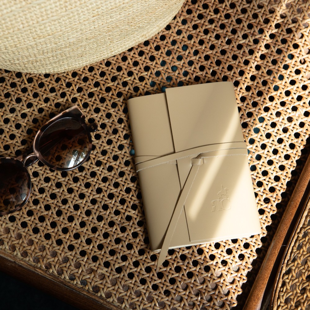 A beige leather-bound notebook with a wraparound tie closure rests on a woven rattan tabletop, illuminated by warm natural sunlight. A pair of dark sunglasses and the brim of a straw hat are partially visible nearby, creating a relaxed and sophisticated travel-inspired setting. The notebook features an embossed monogram on the cover, emphasizing its elegant and refined design.