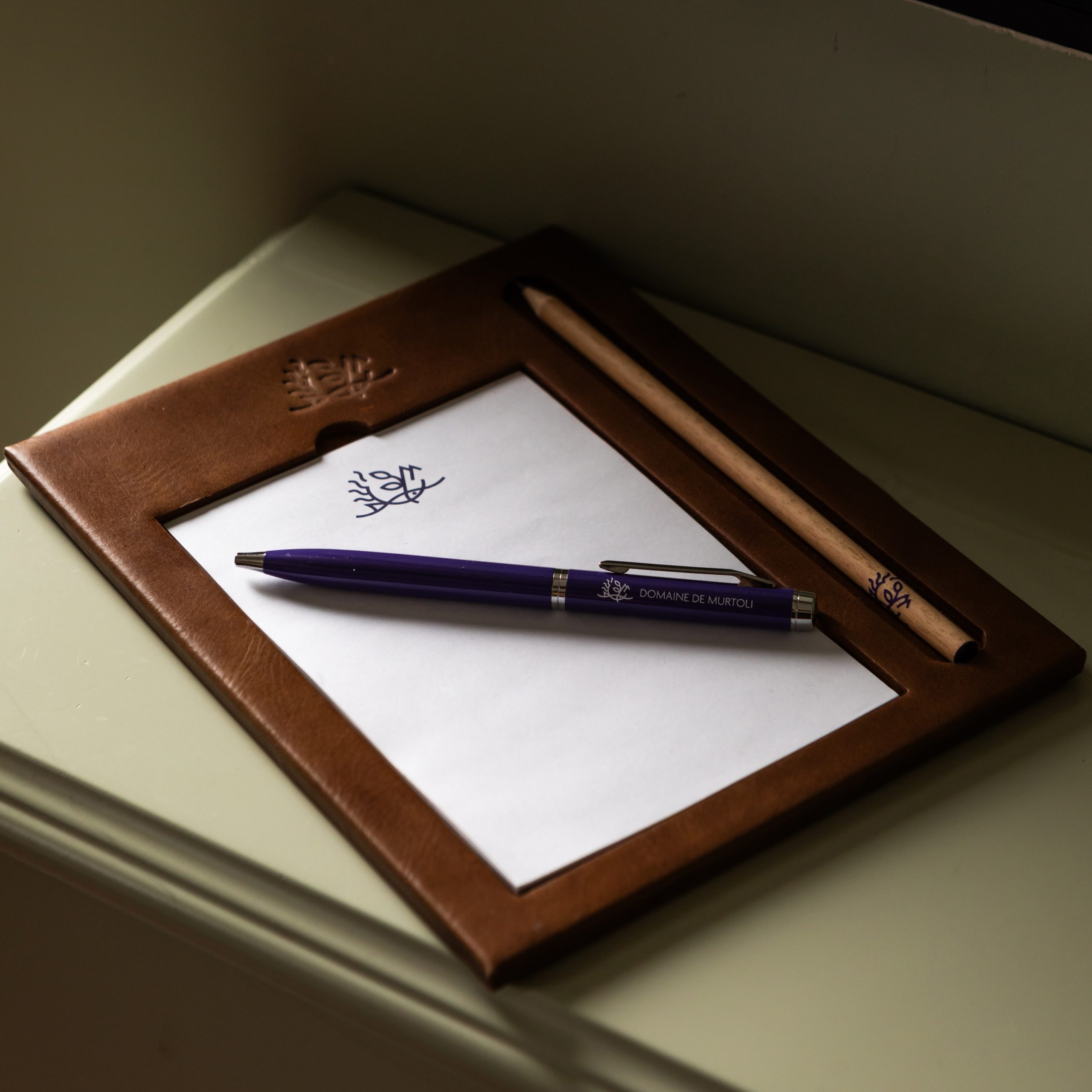 Leather writing pad with a notepad, pencil, and branded pen displayed on a bedside table.