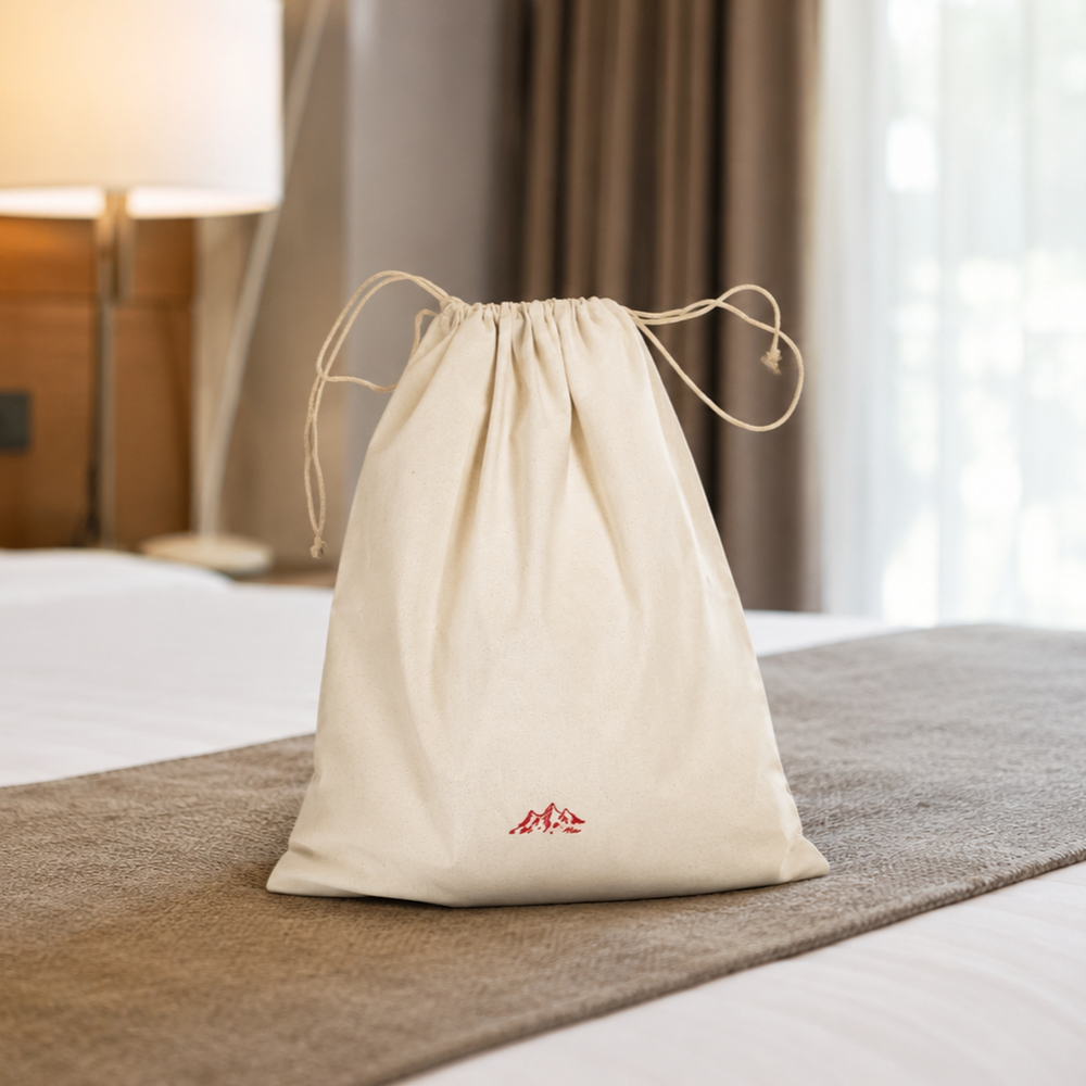 A natural cotton drawstring laundry bag stands upright on a neatly made hotel bed. The bag features a small red mountain logo near the bottom and is placed on a textured bed runner. In the softly blurred background, a bedside lamp, pillows, and curtains create a warm and inviting guest room atmosphere.
