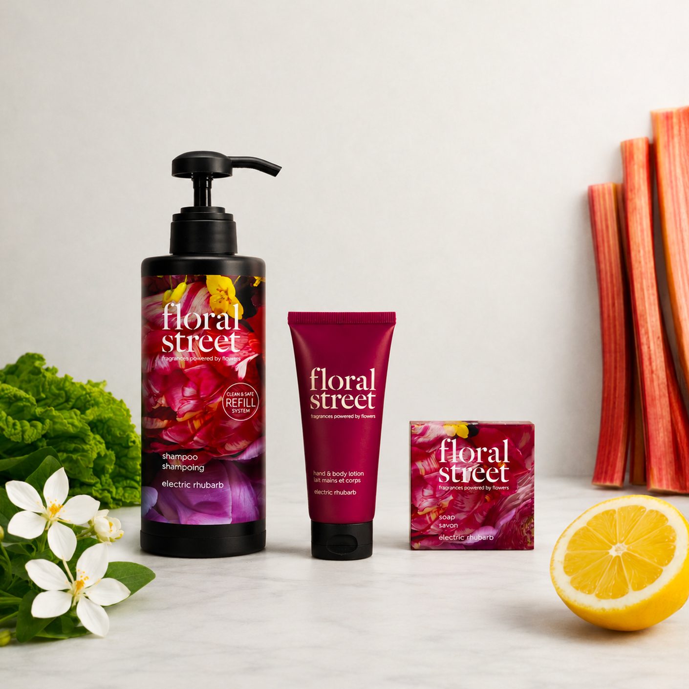 Floral Street Electric Rhubarb bath and body products including refillable shampoo, hand and body lotion, and soap, styled with fresh rhubarb stalks, white flowers, green leaves, and lemon on a clean background.