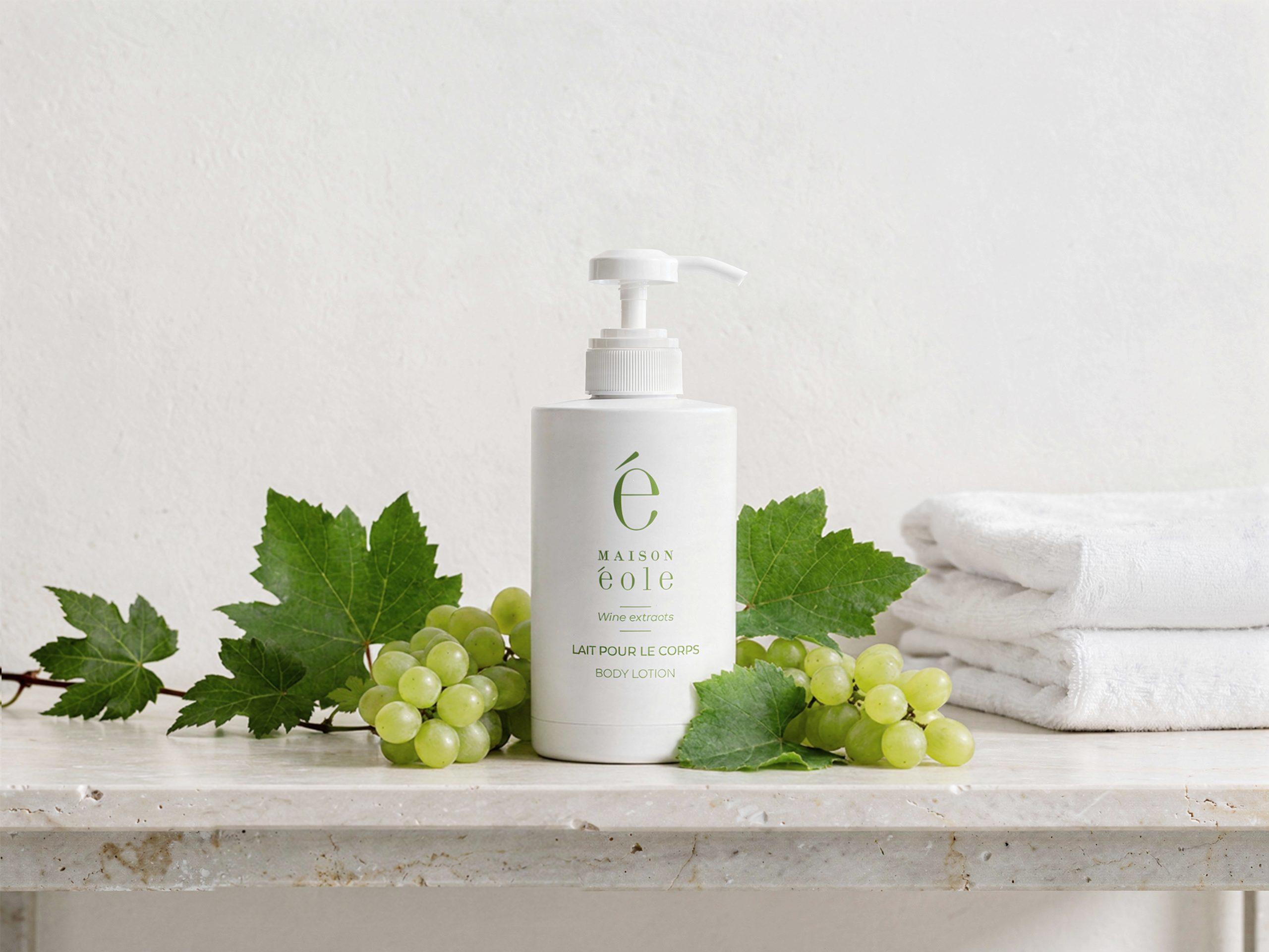 Maison Éole body lotion bottle with pump dispenser placed on a stone countertop, surrounded by green grapes and vine leaves, with folded white towels in the background.