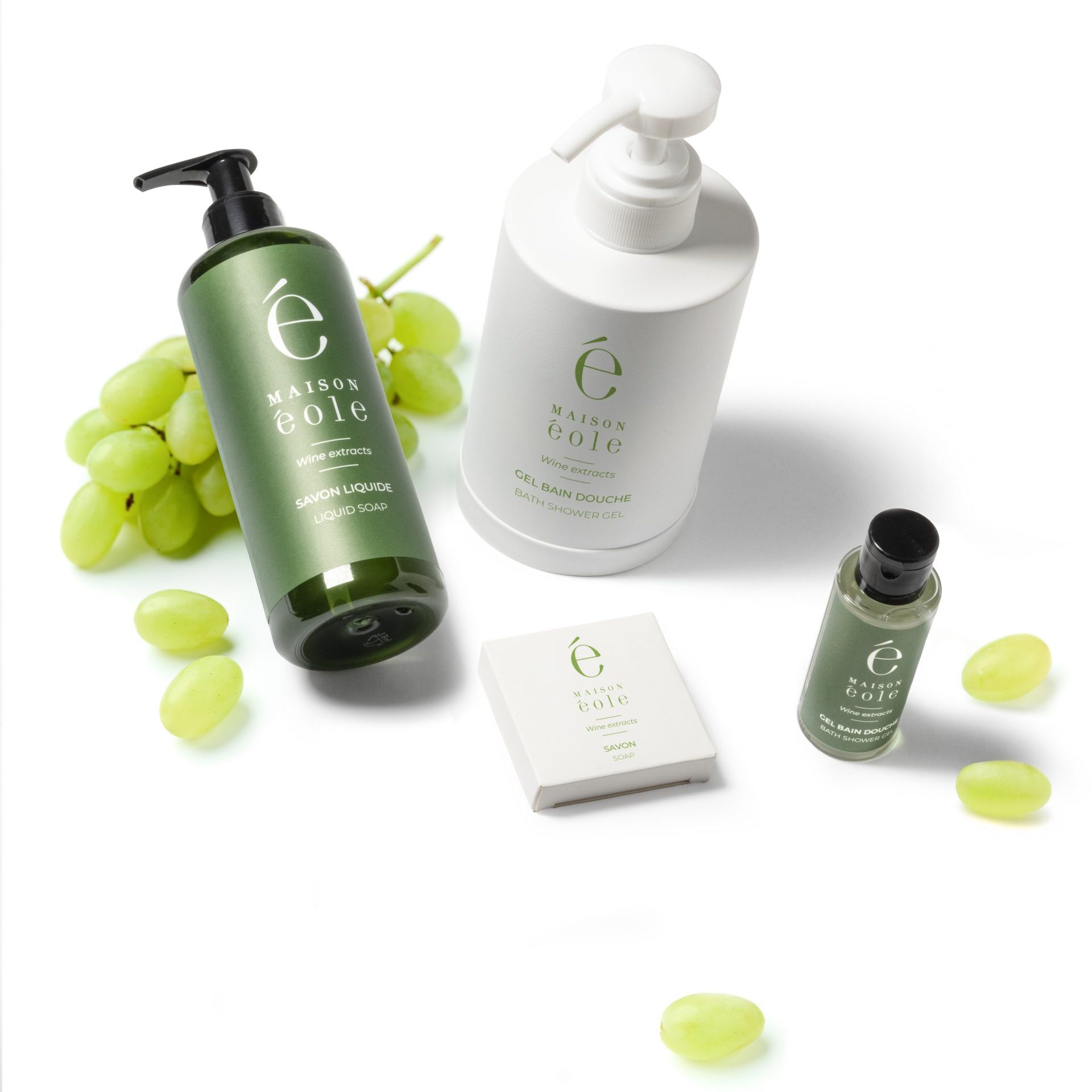 Maison Éole hospitality toiletries collection displayed on a white background, including liquid soap, bath and shower gel, bar soap, and a travel-size bottle, arranged with green grapes.